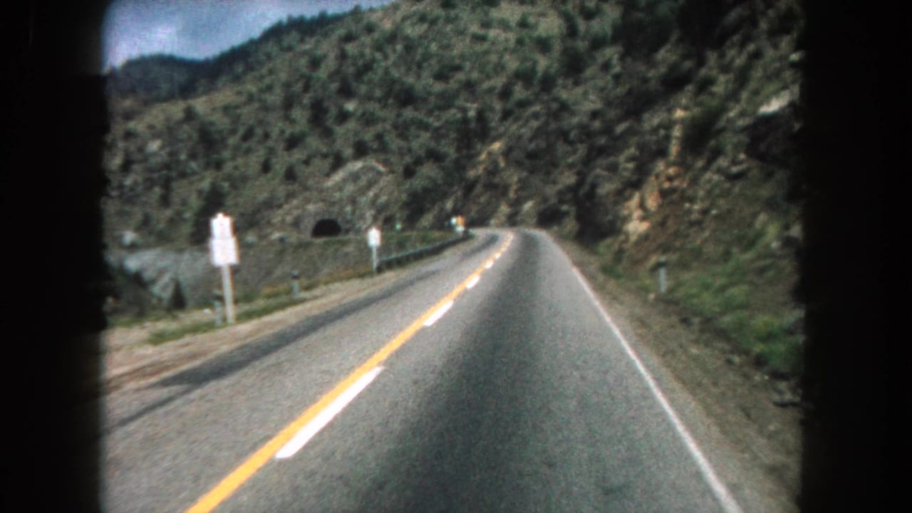 Forward view down a two-lane mountain road with rocky green hillsides and a tunnel ahead.