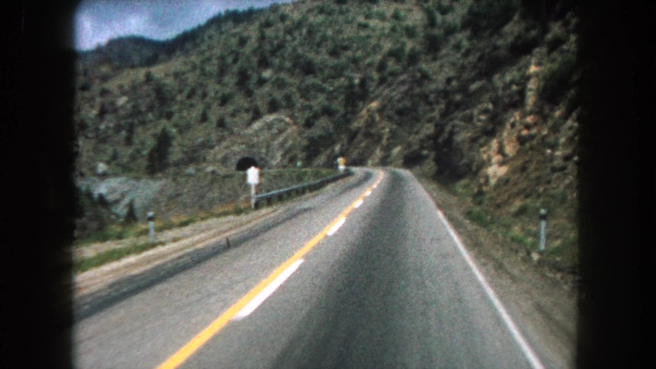 Curving mountain road seen from a moving car, with guardrail, signs, hillside, and tunnel ahead.