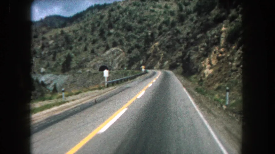 Forward-facing 1962 Summit County, Colorado road view shows a bend in the highway, roadside markers, green hillsides, and a tunnel entrance.