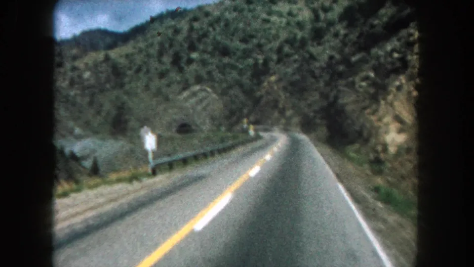 A slightly soft 1962 8mm frame shows a Summit County, Colorado mountain road curving past signs, guardrail, and green rocky terrain.