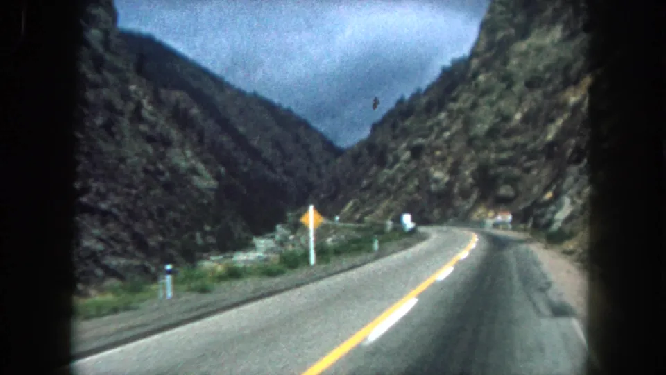 1962 Summit County, Colorado driving view shows a two-lane road curving through a narrow rocky valley with roadside signs and green slopes.