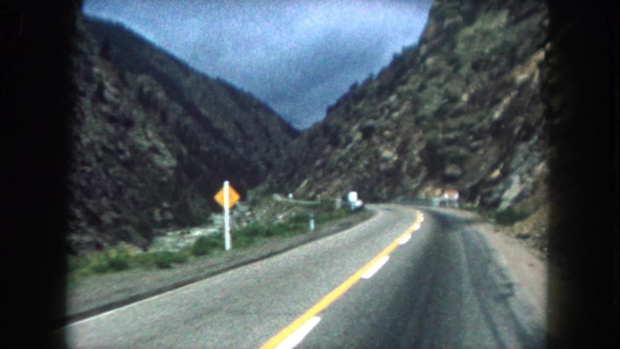 Winding two-lane road through a rocky mountain valley with a yellow warning sign near the curve.
