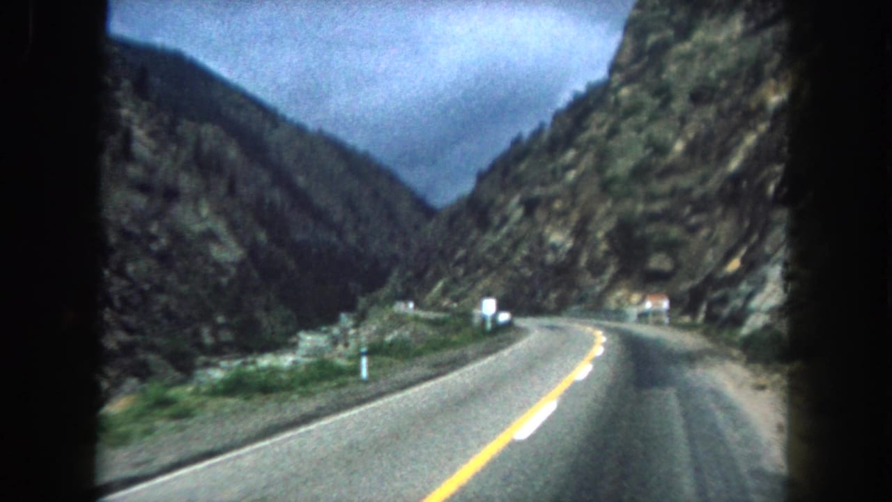 Curving mountain road bordered by rocky cliffs, roadside posts, and cloudy sky.