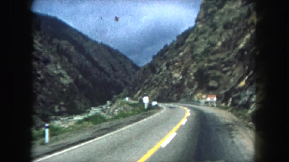1962 Colorado home-movie frame showing a winding Summit County road from inside a moving vehicle, with rocky walls, dark forested slopes, and roadside markers along the curve.