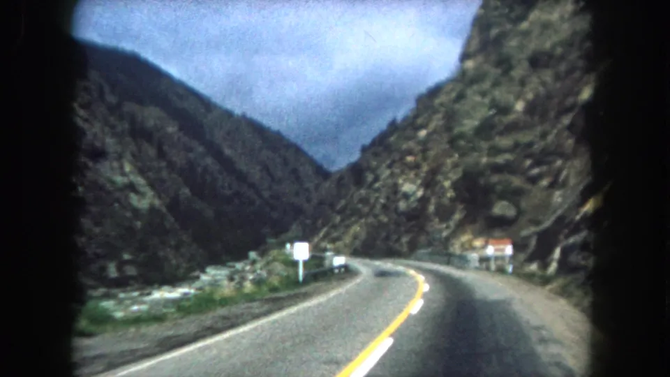 Final sampled 8mm frame from the 1962 Summit County road trip clip, showing an empty mountain highway bending through a rocky valley with roadside signs and a cloudy sky.