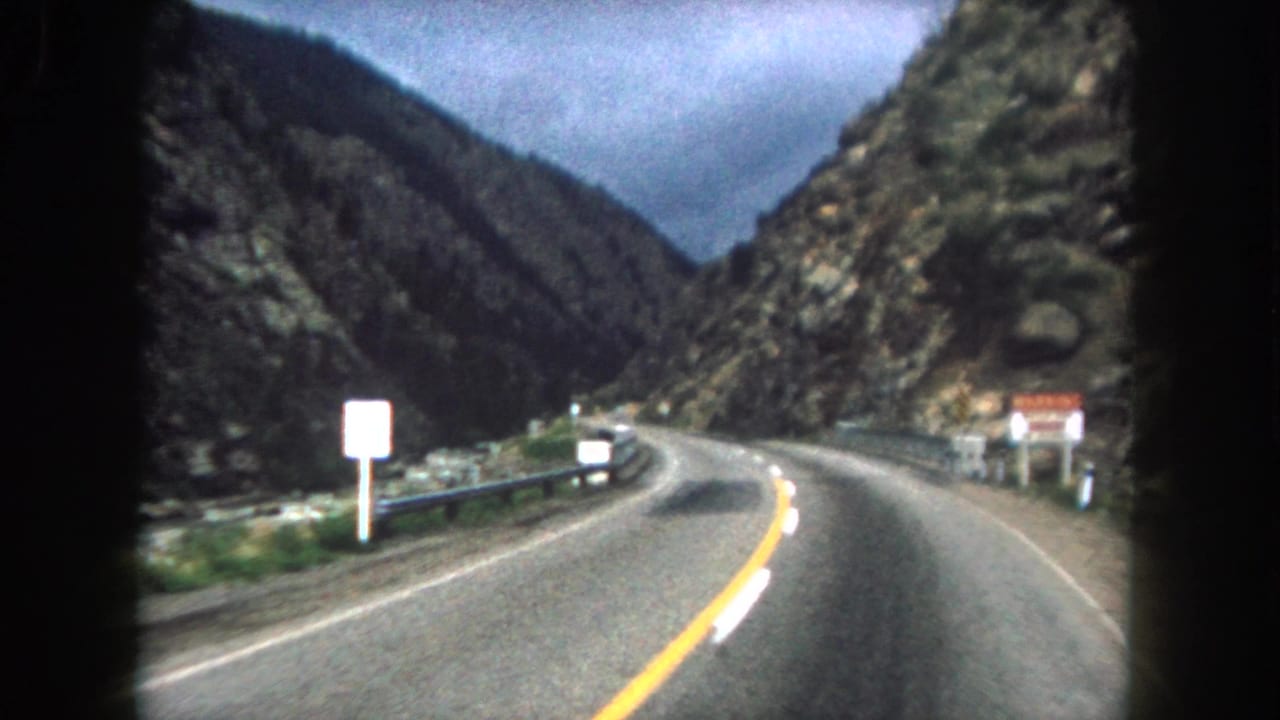 Moving 8mm view along a curving two-lane road in Summit County, Colorado, with guardrail and steep rock faces visible.