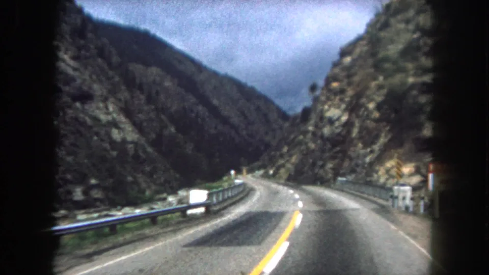 1962 home-movie frame of a two-lane Colorado mountain road descending through rocky terrain from inside a moving car.