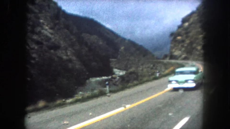 A green period car approaches on the opposite lane of a Summit County mountain road in this 1962 8mm travel frame.