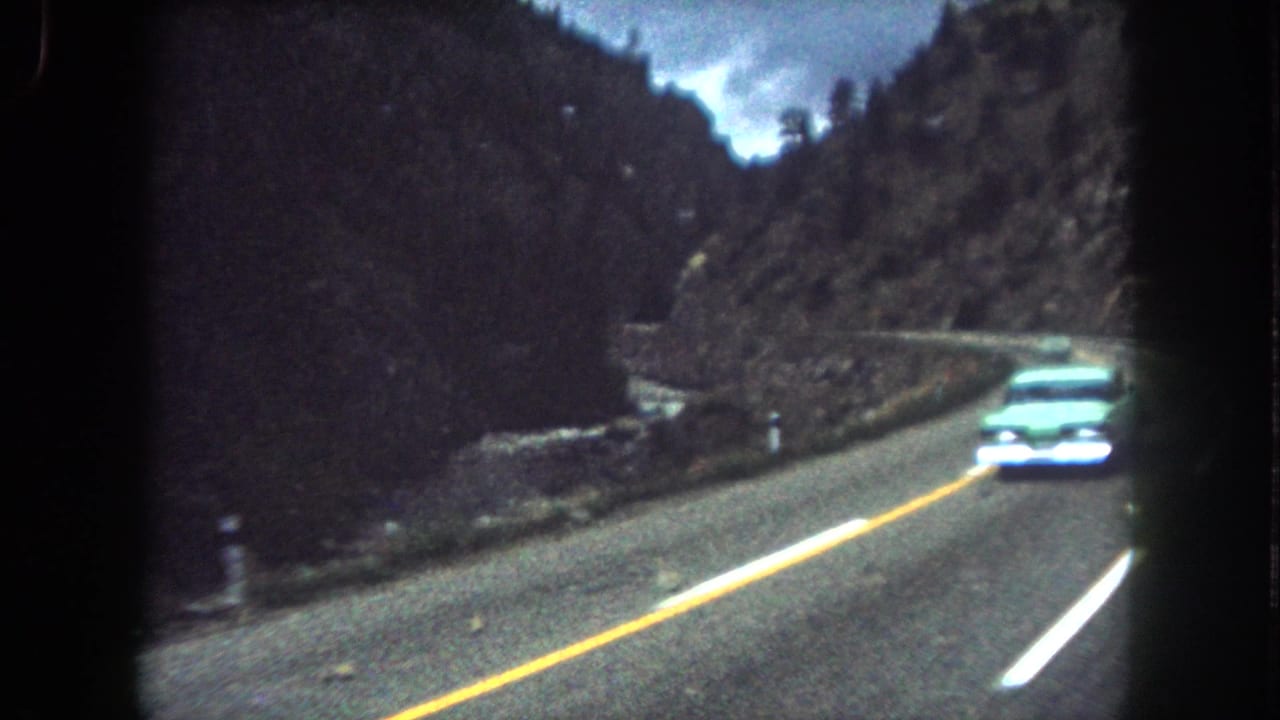 Pale vintage car approaches on a winding mountain road with yellow center line and dark wooded slopes.