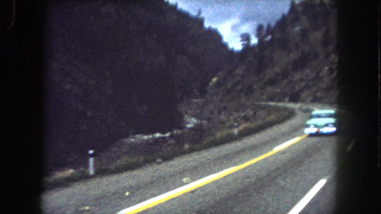 Vintage car rounds a curve on a mountain highway beside rocky terrain and evergreen-covered slopes.