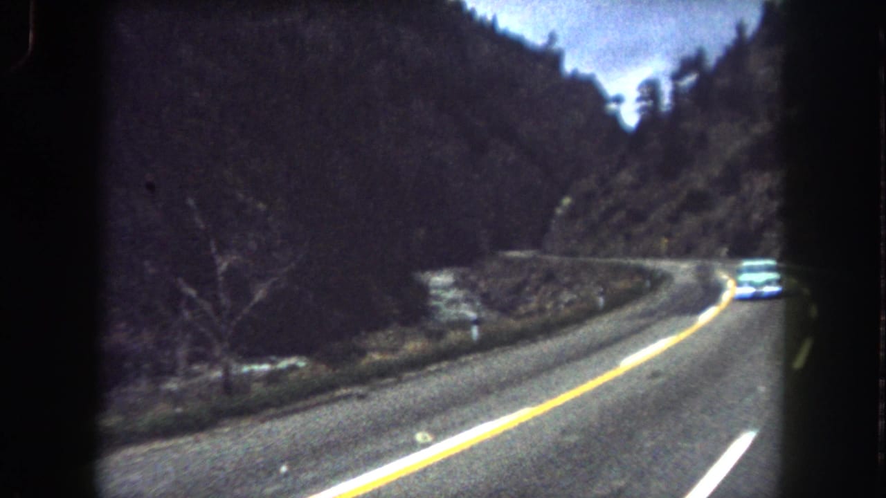 1962 Summit County, Colorado 8mm frame showing a car ahead on a curving mountain road with river and wooded slopes nearby.
