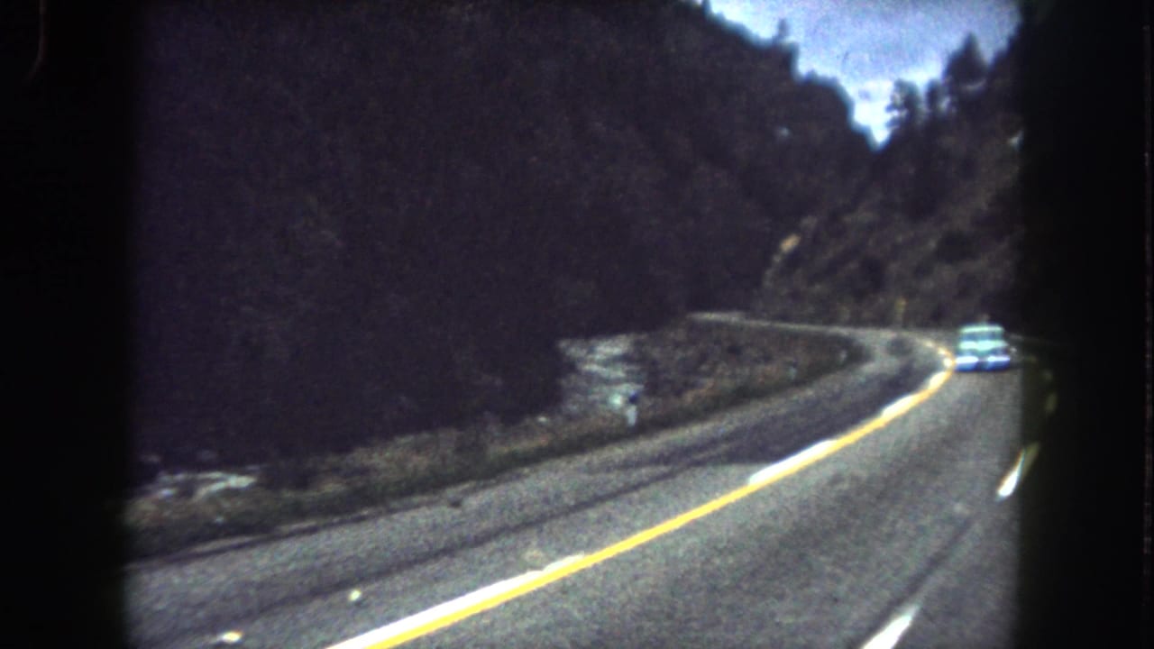 1962 Summit County, Colorado road view with a light-colored car rounding a curve beside a river and forested mountain slope.