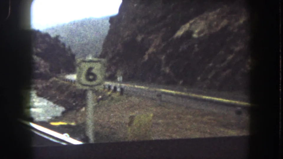 A route marker stands close to the shoulder as the 1962 8mm view continues along a Colorado mountain road.