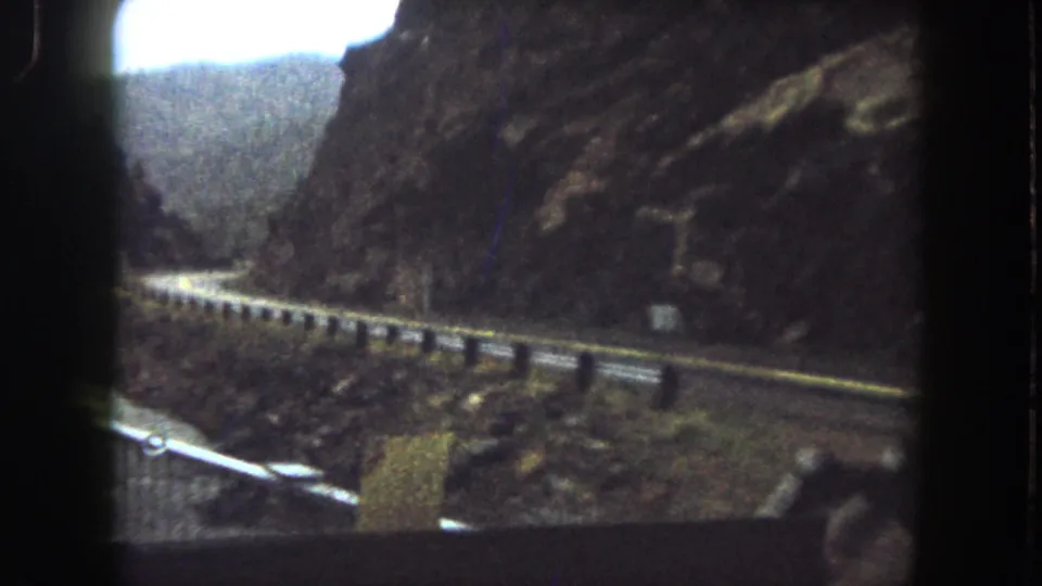 Final frame of a moving 8mm view along a Colorado mountain highway bordered by guardrail, rock walls, and distant hills.