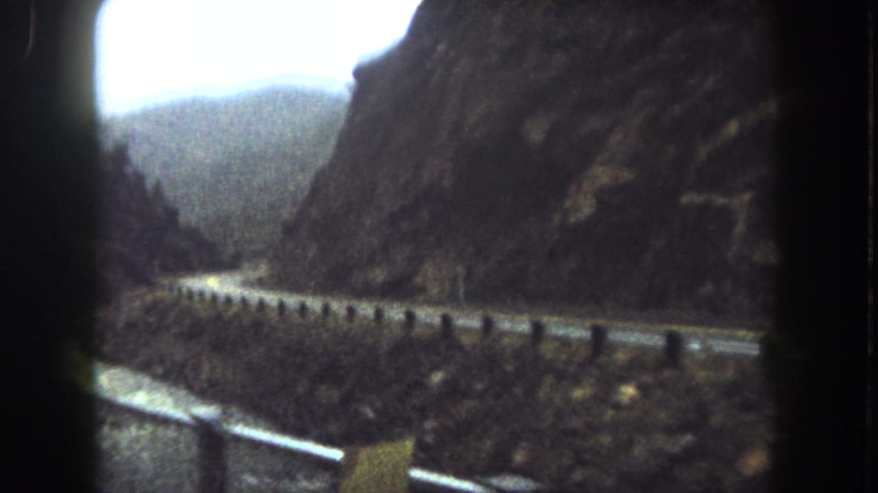 1962 Summit County, Colorado home-movie frame showing a road, guardrail, river and dark rocky mountain slopes.