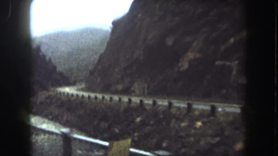 1962 Summit County, Colorado home-movie frame showing a road, guardrail, river and dark rocky mountain slopes.