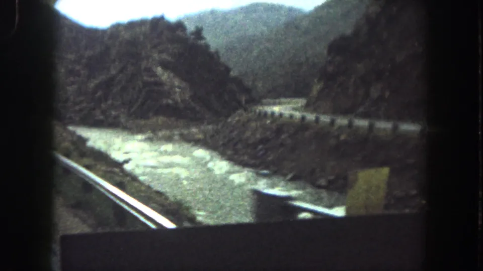 1960s 8mm travel frame showing a river channel, roadside railing and steep Colorado mountain slopes.