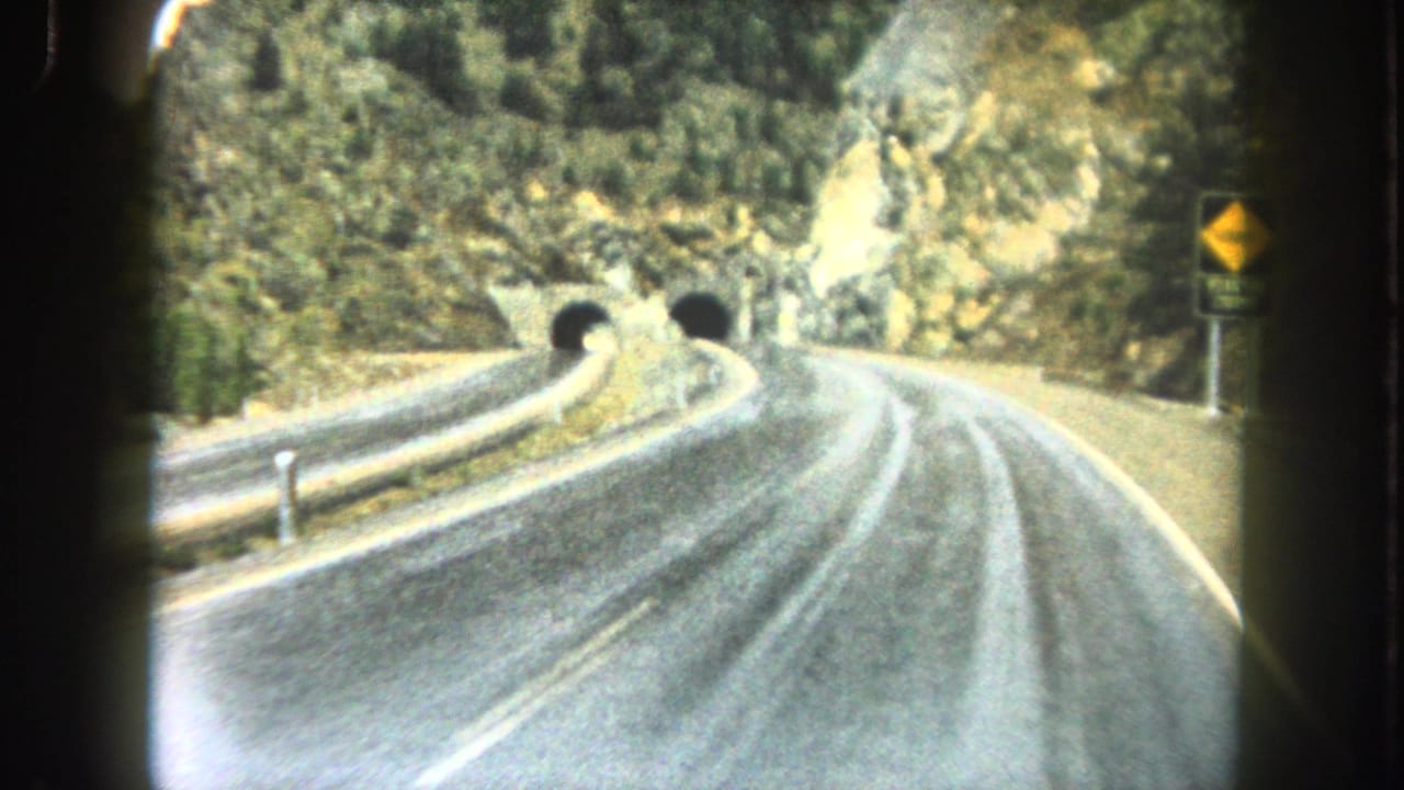 1962 8mm view from a moving vehicle on a wet-looking Summit County mountain road approaching twin tunnel portals.