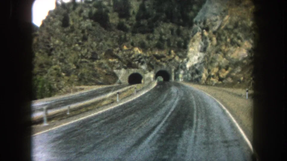 Closer 1962 8mm vehicle perspective on a Summit County mountain road approaching dark tunnel mouths in rock.