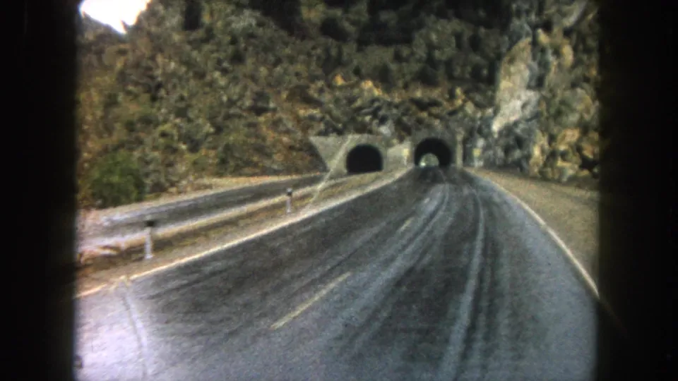 End-frame 8mm view of a Summit County road nearing two tunnel portals set into a rocky, tree-covered slope.