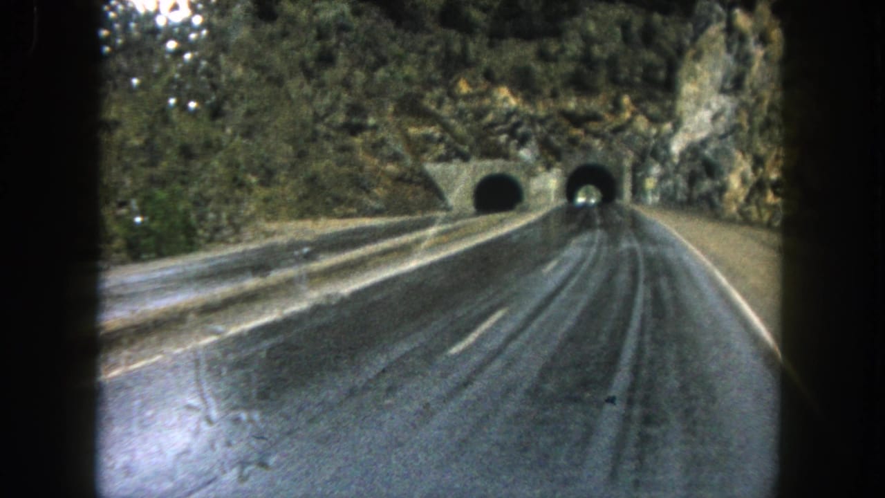 1962 Summit County, Colorado home-movie frame showing a rain-slick highway curving toward two tunnels in a rocky mountainside.