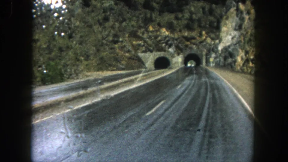 1962 Summit County, Colorado home-movie frame showing a rain-slick highway curving toward two tunnels in a rocky mountainside.