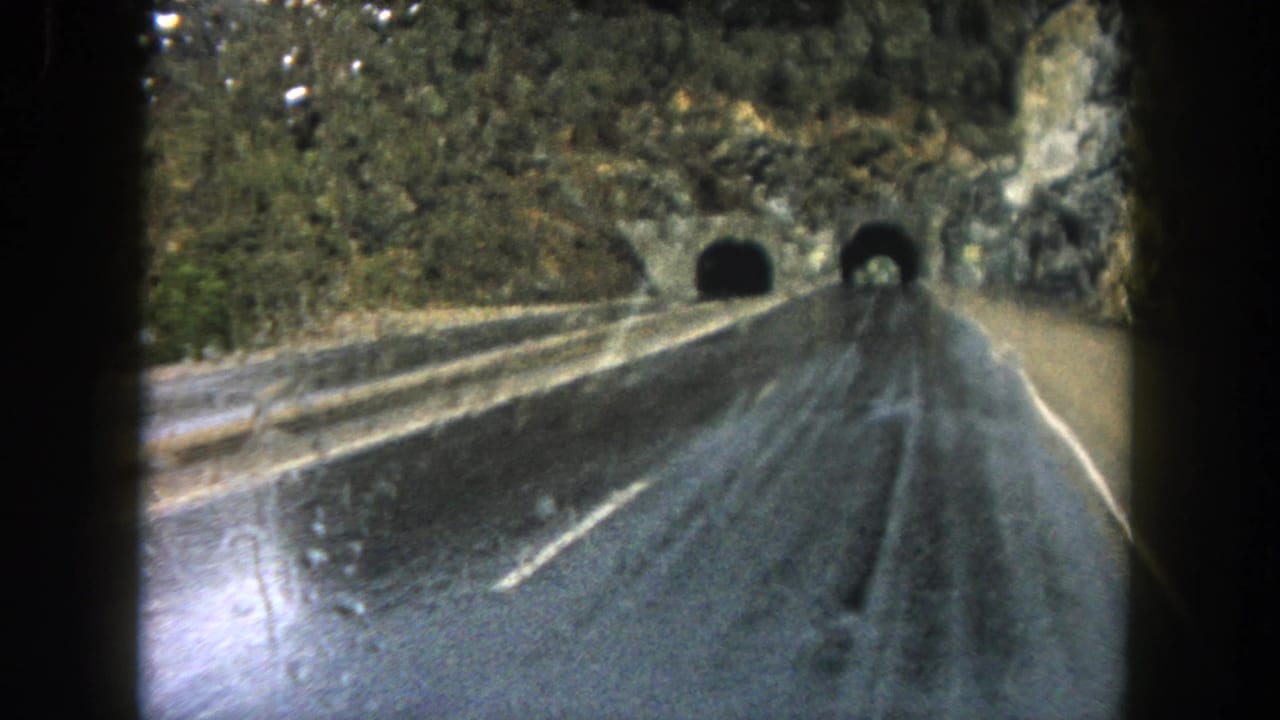 Rainy windshield view of a wet road leading toward dark tunnel entrances in the mountainside.