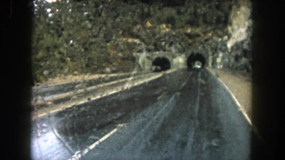 1962 color 8mm view of a wet Summit County mountain road, framed by the vehicle interior as it approaches tunnels cut into rock.