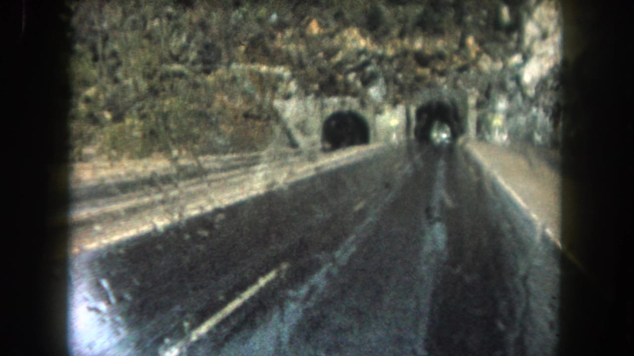 Wet two-lane road approaching tunnel openings in a rocky mountainside.