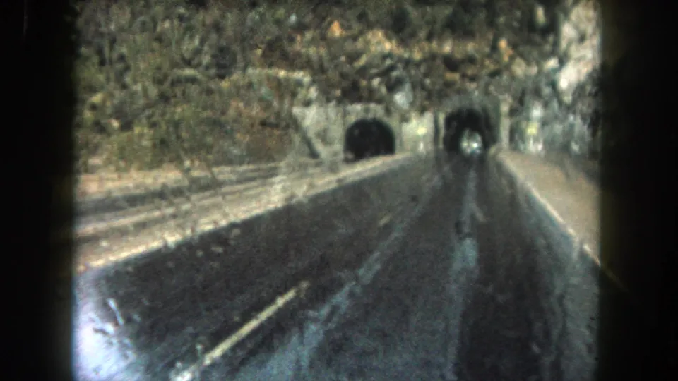 1962 Summit County, Colorado 8mm frame showing a wet highway leading toward dark tunnel portals in a rocky slope.