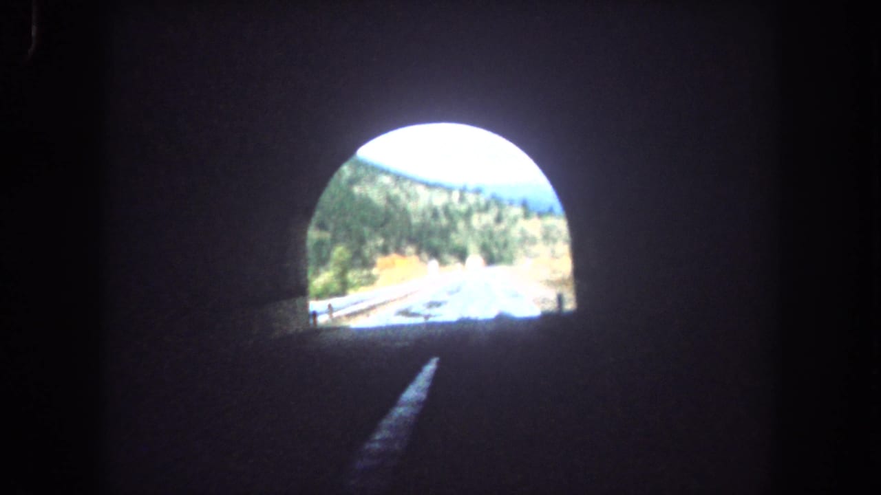 Dark tunnel interior with a bright arched exit revealing a wet mountain road beyond.