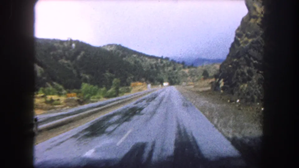 1962 home-movie frame showing a wet Summit County mountain road after leaving the tunnel, with forested hills ahead.
