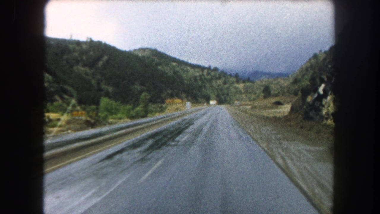 1962 8mm view from a moving vehicle on a wet highway through Summit County, Colorado, with wooded hills ahead.