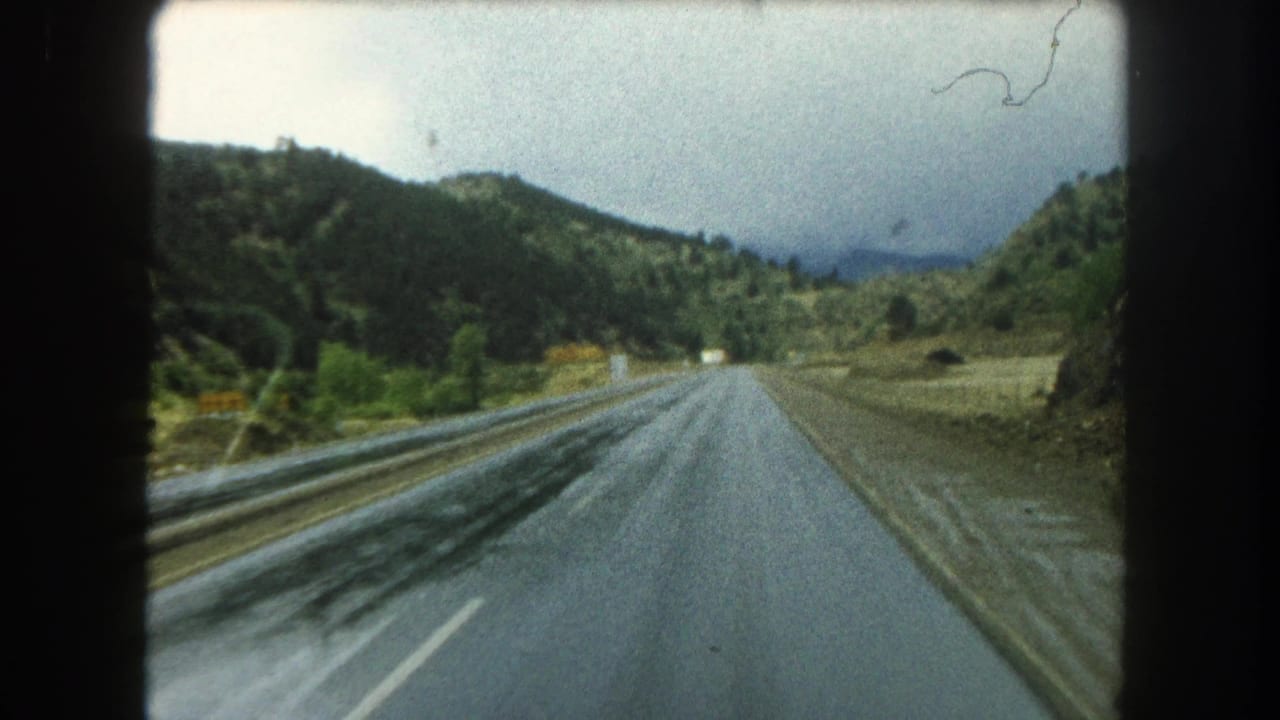 Vehicle-point-of-view frame of a wet Summit County mountain road in 1962, bordered by green slopes and brush.