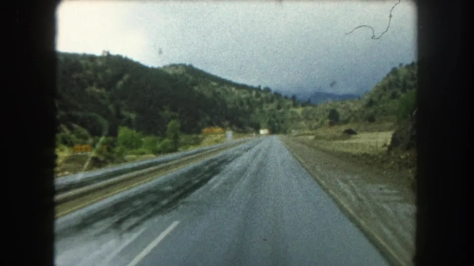 Vehicle-point-of-view frame of a wet Summit County mountain road in 1962, bordered by green slopes and brush.