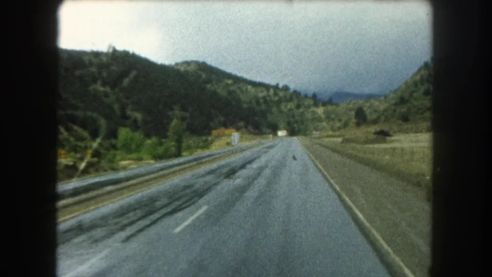 1962 8mm road-travel frame showing a wet two-lane highway in Summit County with trees, hills, and a small roadside sign.