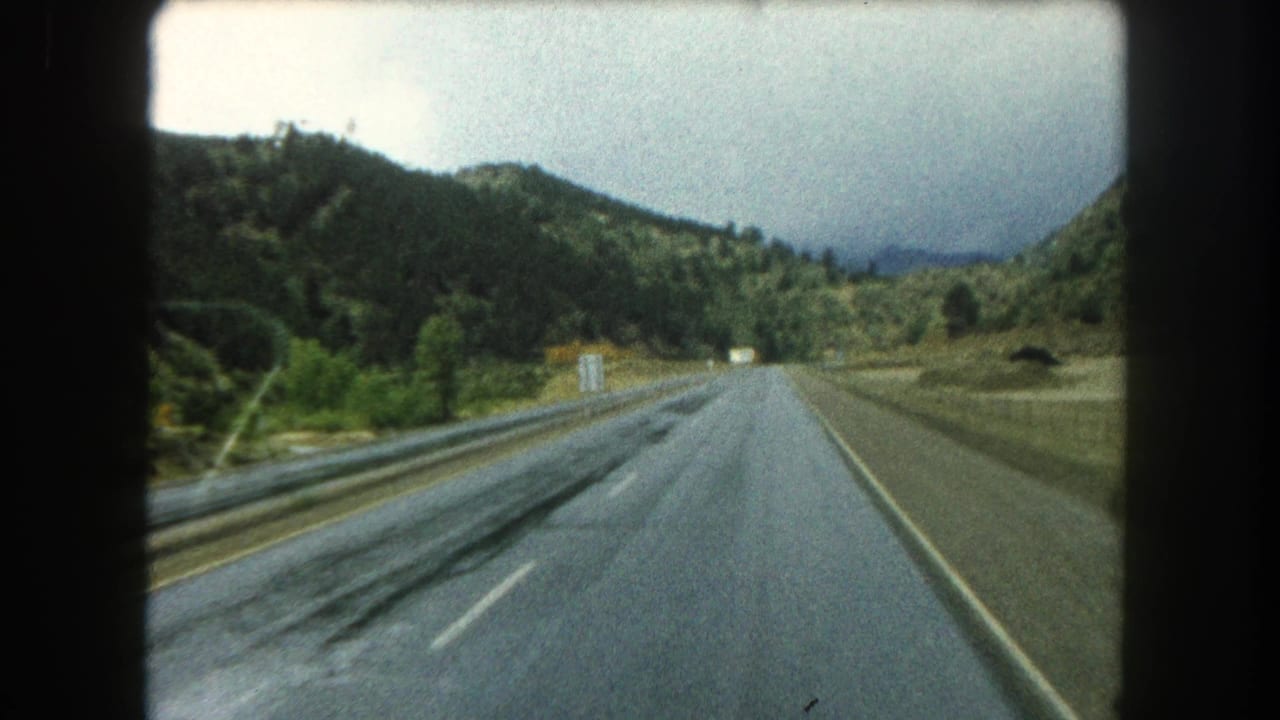 1962 8mm view along a wet highway in Summit County, Colorado, with forested slopes and guardrails ahead.
