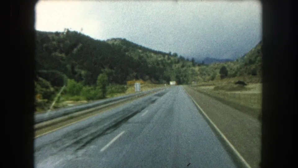 1962 8mm view along a wet highway in Summit County, Colorado, with forested slopes and guardrails ahead.