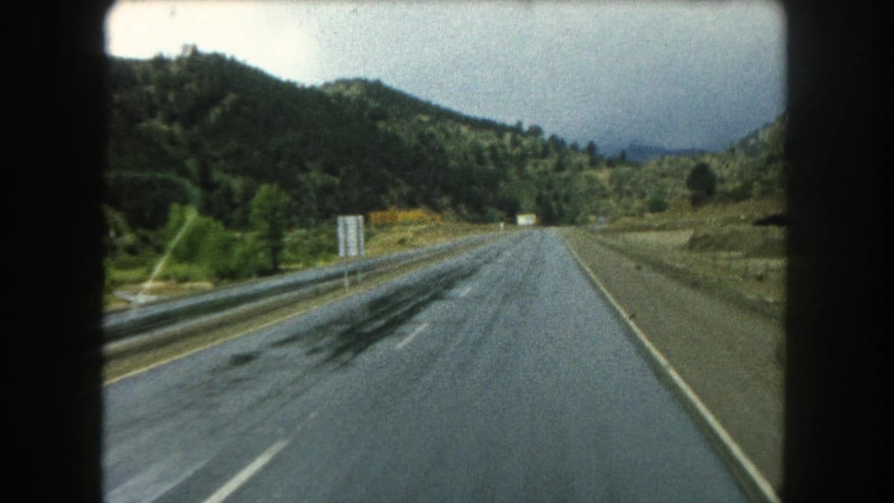 Forward view down a wet highway with guardrails, hillsides, and distant roadside signs.