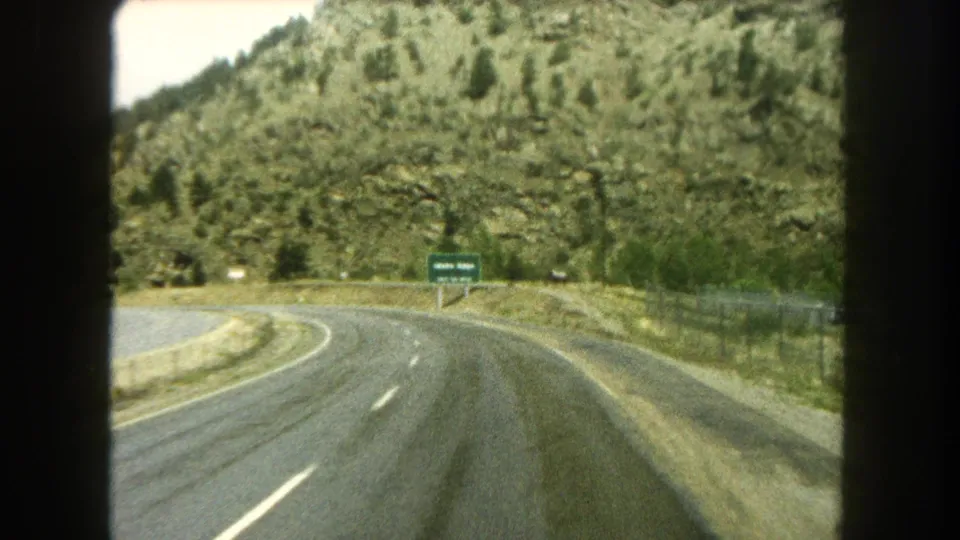 1962 Colorado road-travel frame showing a curve near a green highway sign, fencing, and rocky mountain slope.
