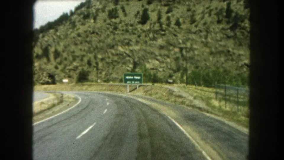 Summit County 8mm frame of a wet highway curve passing a green sign and fence below a scrub-covered slope.