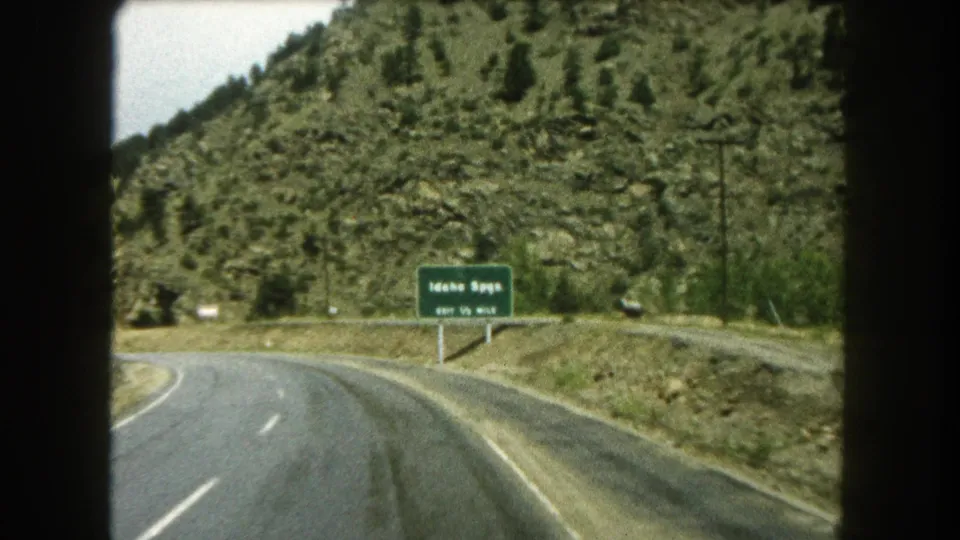 Car-window view of an Idaho Springs exit sign along a curving Colorado mountain road in the 1962 home-movie clip.