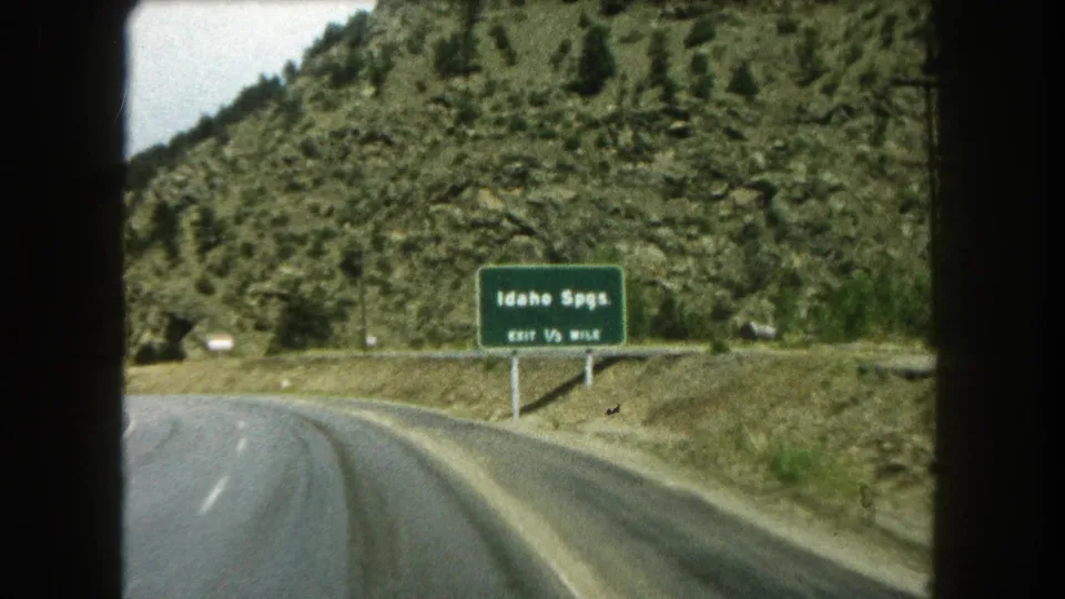 The Idaho Springs exit sign is more legible as the car passes along a Colorado mountain road in 1962 footage.