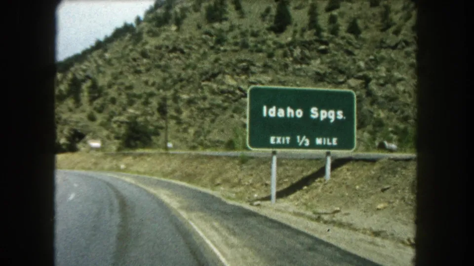 Close 1962 8mm car-view frame of the Idaho Springs sign reading EXIT 1/2 MILE along a Colorado mountain road.