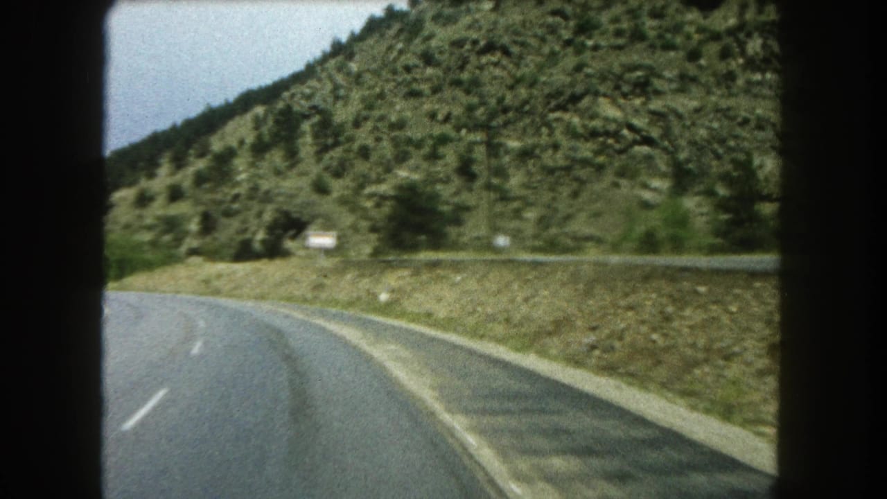 Mountain highway curve with wooded hills and a small roadside sign ahead.