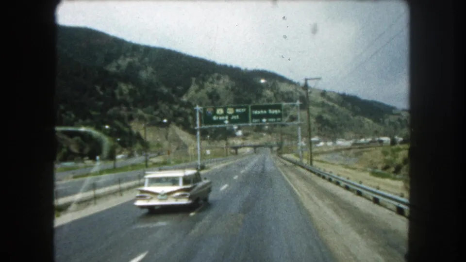1962 8mm frame of a white station wagon approaching overhead signs for Grand Junction and Idaho Springs in Colorado.