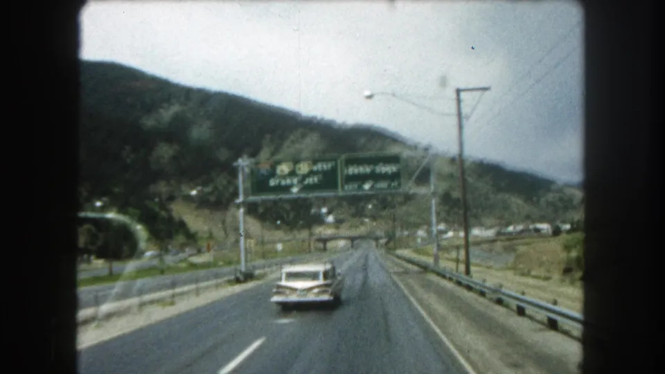 Moving-vehicle view of a white station wagon on a Colorado freeway with overhead signs and mountain hills beyond.