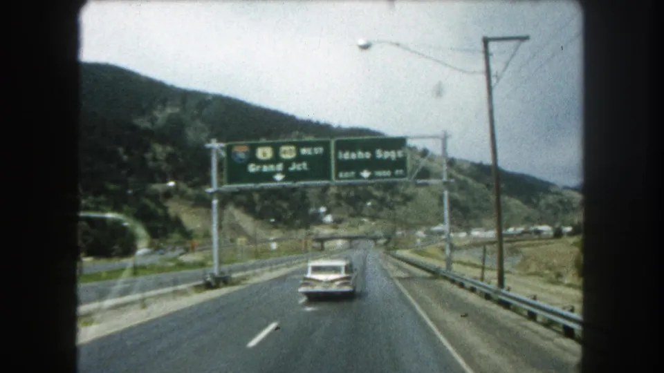 1962 8mm Colorado highway footage showing overhead signs for I-80 and U.S. 40 West, with a white sedan driving through hills.
