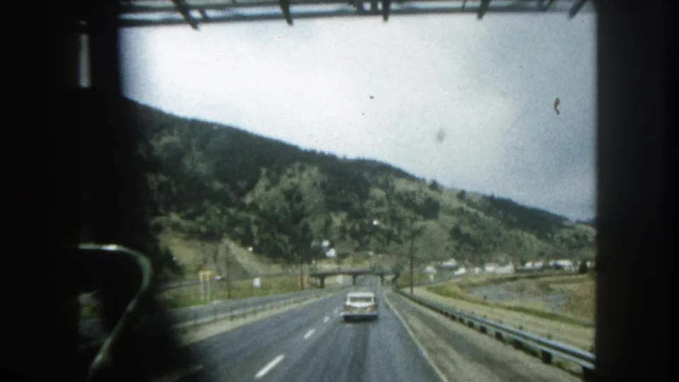 1962 8mm travel view of a Colorado mountain highway, with a white sedan ahead and wooded slopes beyond the road.