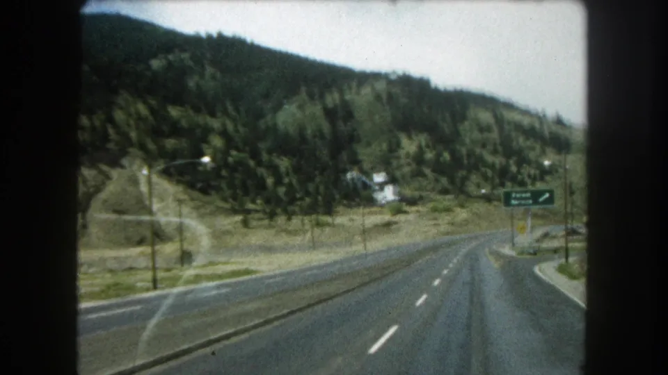 1962 Colorado highway footage showing the roadway bending near an exit sign, with wooded mountain slopes in the background.